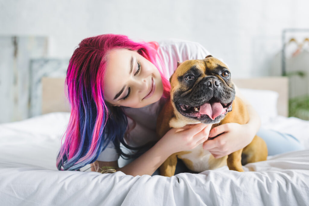 garota com cabelo colorido acariciando e olhando para um lindo buldogue francês enquanto estava deitada na cama